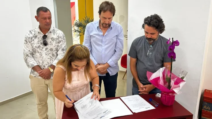 Secret&aacute;ria Mariluci Bilck de vestido rosa claro assina os documentos da reforma do antigo pr&eacute;dio do UCCA sobre um balc&atilde;o vermelho. Ao seu lado, o vice-prefeito Cristiano Costa e um t&eacute;cnico da equipe de engenharia observam o documento. &Agrave; direita, um arranjo de orqu&iacute;deas decora o ambiente.