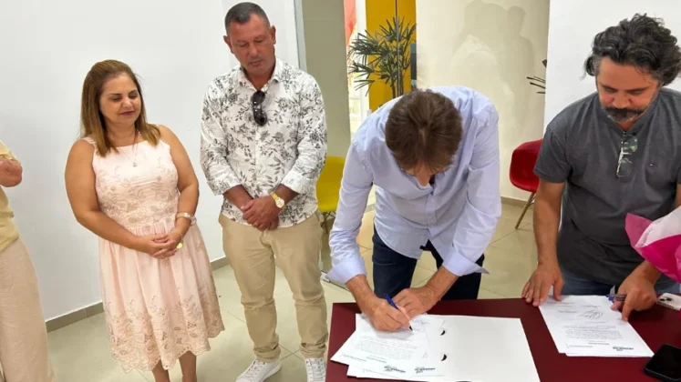 O prefeito de Ararangu&aacute;, C&eacute;sar Cesa, aparece em destaque assinando a Ordem de Servi&ccedil;o. Ele veste uma camisa social azul clara e est&aacute; concentrado no documento sobre o balc&atilde;o. Outras autoridades e familiares do homenageado observam o momento solene ao fundo.