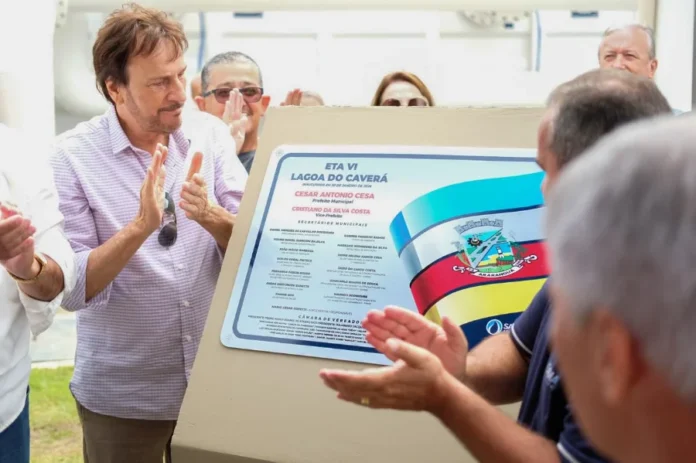 Araranguá inaugura ETA VI e amplia abastecimento de água Autoridades municipais, incluindo o prefeito César Cesa (vestindo camisa xadrez lilás à esquerda), aplaudem sorridentes diante da placa inaugural recém-descerrada da Estação de Tratamento de Água ETA VI - Lagoa do Caverá. A placa exibe o brasão de Araranguá, a data de inauguração e os nomes dos gestores municipais e vereadores. A imagem ilustra o ato oficial de entrega da obra mencionado na matéria.