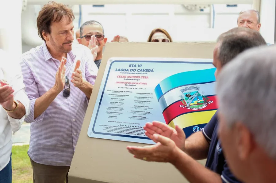Autoridades municipais, incluindo o prefeito César Cesa (vestindo camisa xadrez lilás à esquerda), aplaudem sorridentes diante da placa inaugural recém-descerrada da Estação de Tratamento de Água ETA VI - Lagoa do Caverá. A placa exibe o brasão de Araranguá, a data de inauguração e os nomes dos gestores municipais e vereadores. A imagem ilustra o ato oficial de entrega da obra mencionado na matéria.