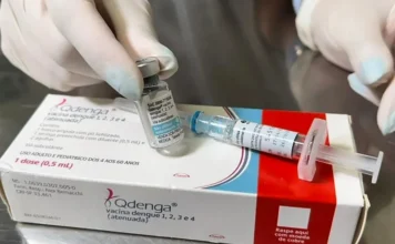 Araranguá inicia vacinação contra dengue para jovens Mãos de um profissional de saúde com luvas seguram um frasco e a caixa da vacina Qdenga contra a dengue, ao lado de uma seringa, ilustrando o imunizante utilizado na campanha de vacinação para crianças e adolescentes em Araranguá.