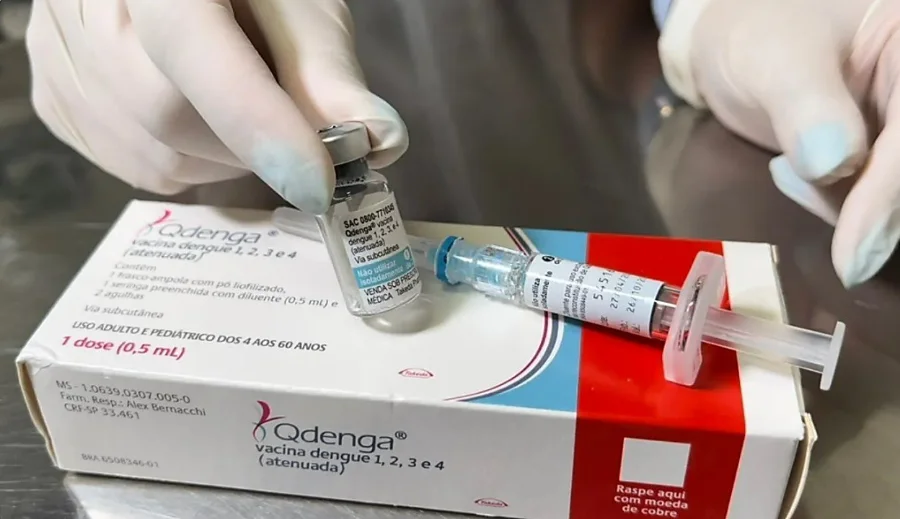Mãos de um profissional de saúde com luvas seguram um frasco e a caixa da vacina Qdenga contra a dengue, ao lado de uma seringa, ilustrando o imunizante utilizado na campanha de vacinação para crianças e adolescentes em Araranguá.
