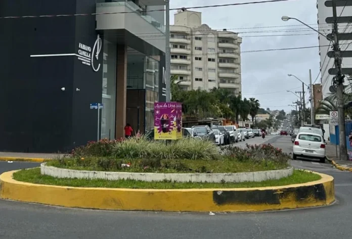 Araranguá retoma obras na Rua Caetano Lummertz nesta quarta Vista da Rua Caetano Lummertz, em Araranguá, sob céu nublado. Em primeiro plano, uma rotatória com canteiro de flores e meio-fio pintado de amarelo. À esquerda, destaca-se um edifício moderno de fachadas preta e branca com o letreiro "Fabiano Canella". Ao longo da via, veem-se carros estacionados e outros edifícios ao fundo. Este é o trecho que receberá as obras de substituição da rede pluvial citadas no texto.