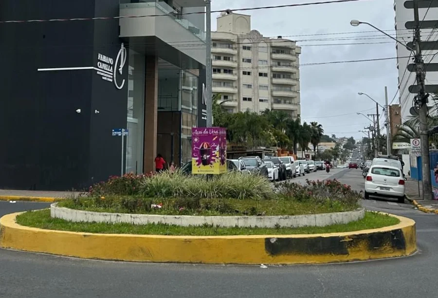Vista da Rua Caetano Lummertz, em Araranguá, sob céu nublado. Em primeiro plano, uma rotatória com canteiro de flores e meio-fio pintado de amarelo. À esquerda, destaca-se um edifício moderno de fachadas preta e branca com o letreiro 