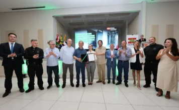 Araranguá terá nova unidade prisional com 686 vagas Autoridades reunidas em palco, com destaque para um homem e uma mulher no centro segurando o documento de ordem de serviço para a construção do novo presídio de Araranguá. Atrás deles, um telão exibe o título do evento e as bandeiras do Brasil e de Santa Catarina. As pessoas ao redor estão aplaudindo ou posando para a foto.