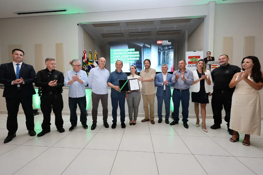 Araranguá terá nova unidade prisional com 686 vagas Autoridades reunidas em palco, com destaque para um homem e uma mulher no centro segurando o documento de ordem de serviço para a construção do novo presídio de Araranguá. Atrás deles, um telão exibe o título do evento e as bandeiras do Brasil e de Santa Catarina. As pessoas ao redor estão aplaudindo ou posando para a foto.