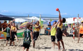 Arena Verão Unesc agita Rincão com esporte e prevenção no mar Atletas disputam a bola no ar durante uma partida intensa do Campeonato de Handebol de Areia na Arena Verão Unesc, em Balneário Rincão. A imagem, sob sol forte, ilustra a alta competitividade do evento esportivo descrito na matéria, com a praia e guarda-sóis ao fundo.
