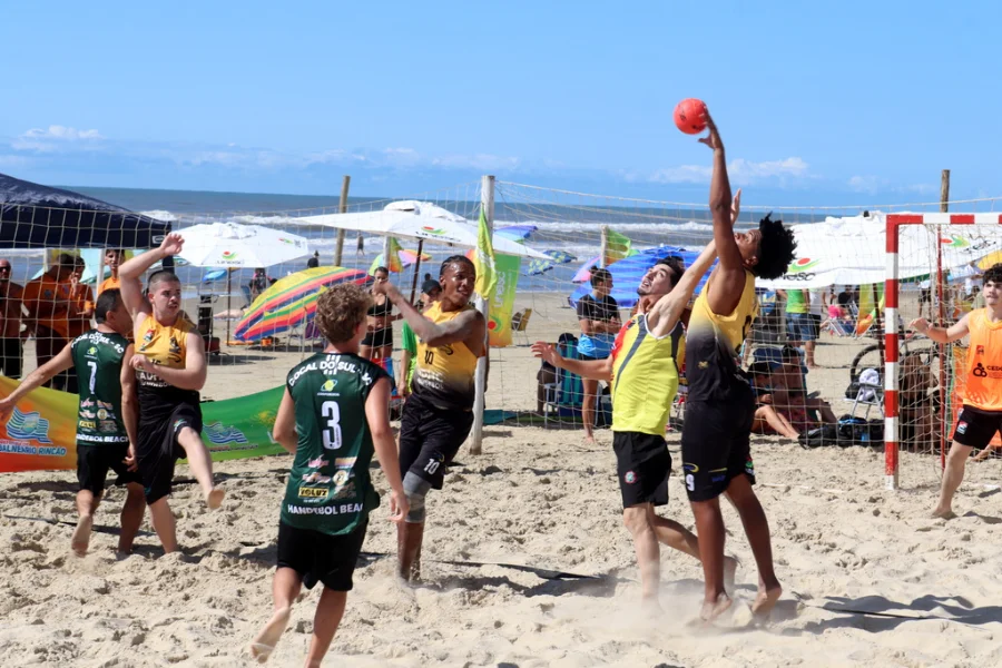 Atletas disputam a bola no ar durante uma partida intensa do Campeonato de Handebol de Areia na Arena Verão Unesc, em Balneário Rincão. A imagem, sob sol forte, ilustra a alta competitividade do evento esportivo descrito na matéria, com a praia e guarda-sóis ao fundo.