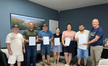 Arroio do Silva autoriza ampliação da EMEB Jardim Atlântico Sete pessoas, entre homens e mulheres, posam em um escritório segurando documentos da ordem de serviço. Na parede azul ao fundo, há um quadro panorâmico da cidade de Balneário Arroio do Silva.