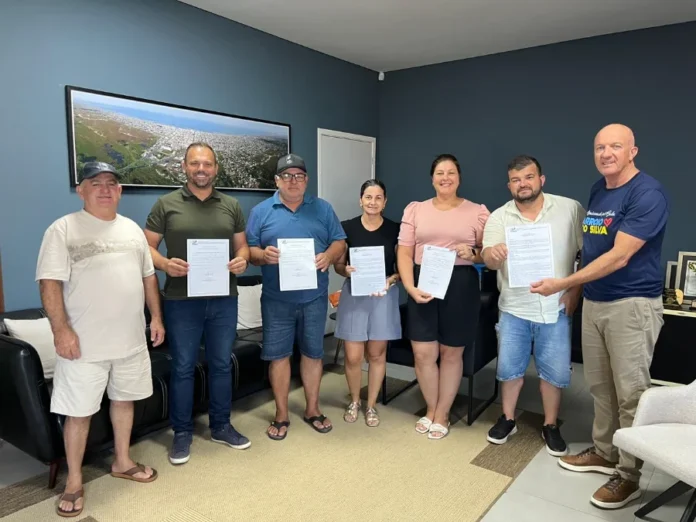 Arroio do Silva autoriza ampliação da EMEB Jardim Atlântico Sete pessoas, entre homens e mulheres, posam em um escritório segurando documentos da ordem de serviço. Na parede azul ao fundo, há um quadro panorâmico da cidade de Balneário Arroio do Silva.
