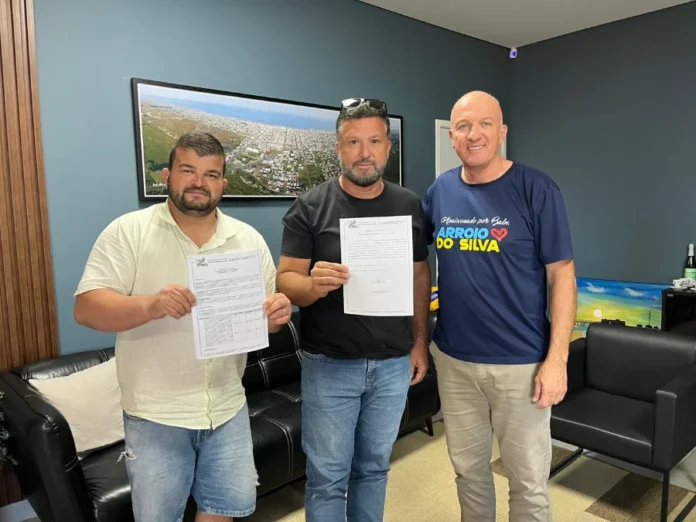 Arroio do Silva autoriza pavimentação na Rua Ermo Fotografia de três homens em pé em um escritório com paredes azuis. À direita, o prefeito Evandro Scaini, vestindo camiseta azul com a escrita "Arroio do Silva", sorri ao lado de dois homens que seguram os documentos da ordem de serviço. Ao fundo, um quadro com a foto aérea da cidade. Ilustra a autorização para pavimentação da Rua Ermo.