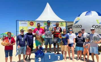 Arroio do Silva realiza 1º Torneio de Arremesso de Tarrafa Fotografia colorida ao ar livre, sob céu azul e ensolarado, mostrando a cerimônia de premiação do 1º Torneio de Arremesso de Tarrafa na beira-mar de Balneário Arroio do Silva. Ao centro, três homens ocupam um pódio azul de madeira; o vencedor (1º lugar) está no degrau mais alto segurando um troféu dourado, ladeado pelos competidores que conquistaram o 2º e 3º lugares. Todos vestem trajes casuais de verão, como bermudas e camisetas.