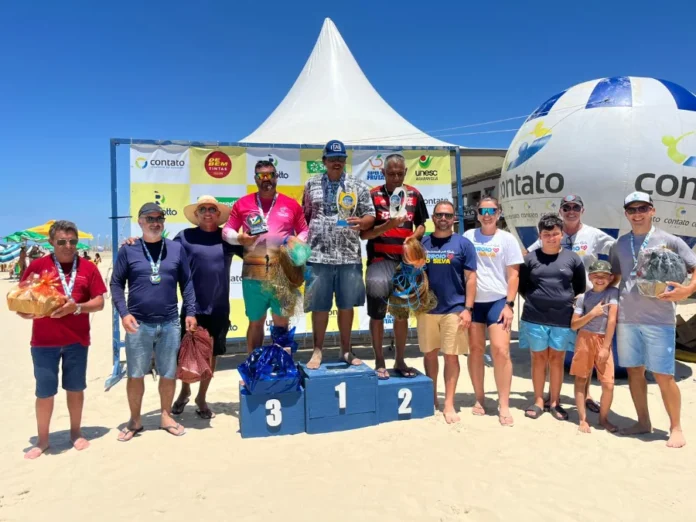 Arroio do Silva realiza 1º Torneio de Arremesso de Tarrafa Fotografia colorida ao ar livre, sob céu azul e ensolarado, mostrando a cerimônia de premiação do 1º Torneio de Arremesso de Tarrafa na beira-mar de Balneário Arroio do Silva. Ao centro, três homens ocupam um pódio azul de madeira; o vencedor (1º lugar) está no degrau mais alto segurando um troféu dourado, ladeado pelos competidores que conquistaram o 2º e 3º lugares. Todos vestem trajes casuais de verão, como bermudas e camisetas.