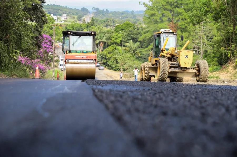 Máquinas pesadas, incluindo um rolo compactador e uma motoniveladora, trabalham na aplicação da nova camada de asfalto na Rua Pedro Dal Toé, em Criciúma. Em primeiro plano, é possível ver o detalhe da textura do asfalto fresco, enquanto ao fundo a via se estende entre áreas de vegetação e residências do bairro Morro Estevão.