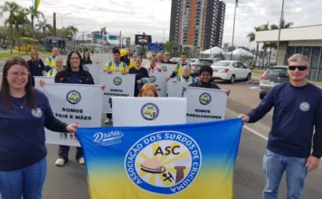 Associação dos Surdos de Criciúma inicia ações em 2026 Fotografia colorida mostra integrantes da Associação dos Surdos de Criciúma (ASC) reunidos em uma via pública durante o dia. Em primeiro plano, uma mulher à esquerda e um homem à direita seguram uma grande bandeira azul e amarela com o logotipo da ASC ao centro. Atrás deles, outras pessoas seguram cartazes brancos com os dizeres: "Somos pais e mães", "Somos professores" e "Somos trabalhadores". O grupo sorri para a câmera, simbolizando a visibilidade e a ocupação de espaços pela comunidade surda na sociedade.