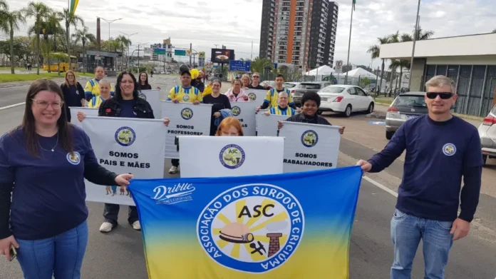 Associação dos Surdos de Criciúma inicia ações em 2026 Fotografia colorida mostra integrantes da Associação dos Surdos de Criciúma (ASC) reunidos em uma via pública durante o dia. Em primeiro plano, uma mulher à esquerda e um homem à direita seguram uma grande bandeira azul e amarela com o logotipo da ASC ao centro. Atrás deles, outras pessoas seguram cartazes brancos com os dizeres: "Somos pais e mães", "Somos professores" e "Somos trabalhadores". O grupo sorri para a câmera, simbolizando a visibilidade e a ocupação de espaços pela comunidade surda na sociedade.