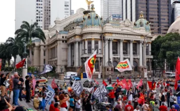 Ato na Cinelândia marca três anos do 8 de janeiro Manifestantes segurando diversas bandeiras, incluindo vermelhas e de centrais sindicais como CUT e CTB, estão reunidos em frente ao histórico edifício do Theatro Municipal, na região da Cinelândia, no centro do Rio de Janeiro, durante um ato público durante o dia.