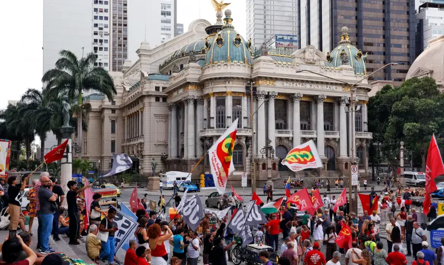 Ato na Cinelândia marca três anos do 8 de janeiro Manifestantes segurando diversas bandeiras, incluindo vermelhas e de centrais sindicais como CUT e CTB, estão reunidos em frente ao histórico edifício do Theatro Municipal, na região da Cinelândia, no centro do Rio de Janeiro, durante um ato público durante o dia.