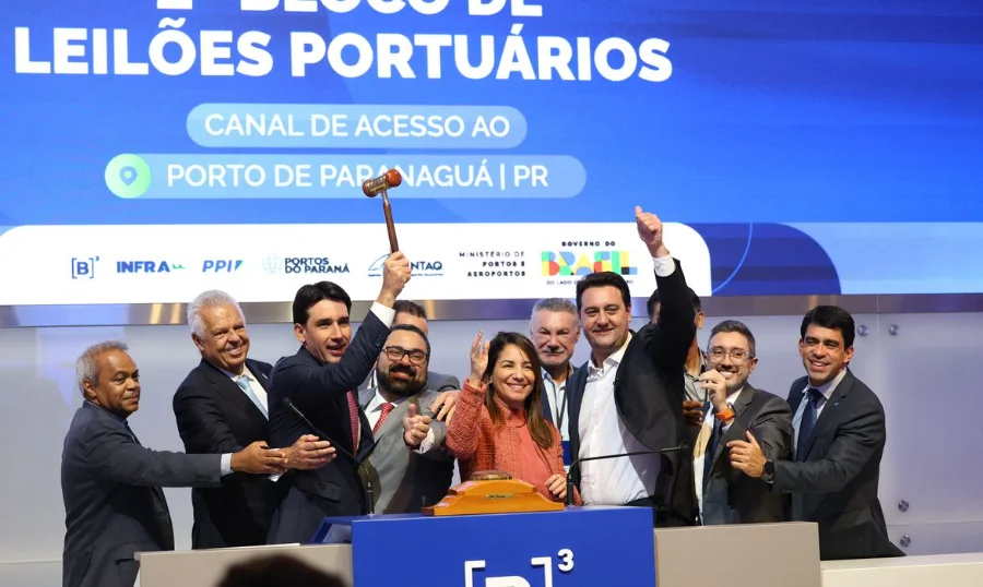 Grupo de autoridades e executivos celebra no palco da B3 durante o leilão do canal de acesso ao Porto de Paranaguá. No centro da imagem, um homem ergue o martelo de madeira (gavel) simbolizando a conclusão do lance, enquanto outros sorriem e fazem sinal de 