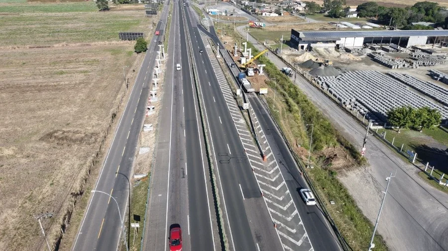 Vista aérea da rodovia BR-101 no trecho de Maracajá (SC), exibindo as frentes de trabalho das novas passarelas nos km 401 e 402. A imagem mostra os pilares de sustentação já instalados às margens das pistas, evidenciando o local onde as vigas serão içadas pela ViaCosteira.