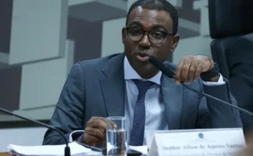 Banco Master tinha só R$ 4 milhões em caixa, diz diretor Fotografia de Ailton de Aquino Santos, diretor de Fiscalização do Banco Central, durante um pronunciamento oficial. Ele é um homem negro, usa óculos de armação escura, terno cinza e gravata azul. Está posicionado atrás de uma bancada com um microfone à sua frente, representando o momento de seu depoimento sobre as irregularidades financeiras do Banco Master.