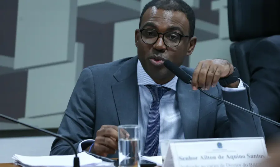 Banco Master tinha só R$ 4 milhões em caixa, diz diretor Fotografia de Ailton de Aquino Santos, diretor de Fiscalização do Banco Central, durante um pronunciamento oficial. Ele é um homem negro, usa óculos de armação escura, terno cinza e gravata azul. Está posicionado atrás de uma bancada com um microfone à sua frente, representando o momento de seu depoimento sobre as irregularidades financeiras do Banco Master.