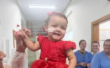 Bebê recebe alta em Criciúma após 400 dias internada A fotografia registra o momento da alta de Ana Lara no Hospital Materno-Infantil Santa Catarina (Hmisc). No centro da imagem, a bebê, uma menina de um ano com cabelos escuros e pequenos laços vermelhos, sorri alegremente enquanto veste um vestido de festa vermelho com detalhes em pedraria. Ela está sendo segurada no colo por um adulto, cujos braços aparecem nas laterais da foto. Ao fundo, três profissionais de saúde em uniformes azuis (scrubs) sorriem, celebrando a recuperação da paciente no corredor da unidade hospitalar.