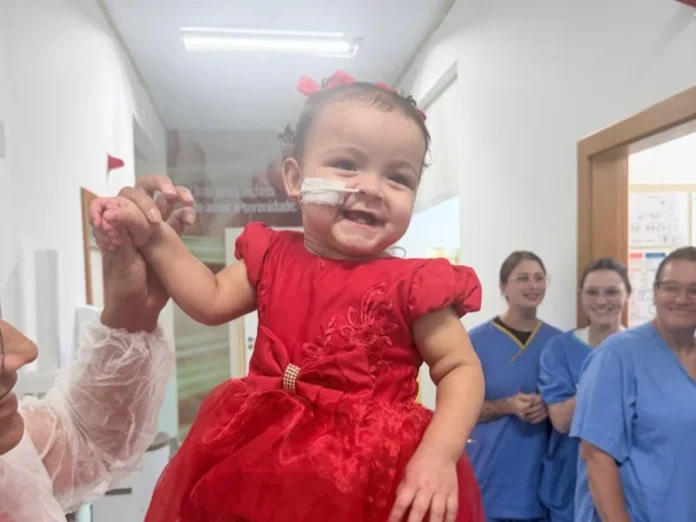 Bebê recebe alta em Criciúma após 400 dias internada A fotografia registra o momento da alta de Ana Lara no Hospital Materno-Infantil Santa Catarina (Hmisc). No centro da imagem, a bebê, uma menina de um ano com cabelos escuros e pequenos laços vermelhos, sorri alegremente enquanto veste um vestido de festa vermelho com detalhes em pedraria. Ela está sendo segurada no colo por um adulto, cujos braços aparecem nas laterais da foto. Ao fundo, três profissionais de saúde em uniformes azuis (scrubs) sorriem, celebrando a recuperação da paciente no corredor da unidade hospitalar.