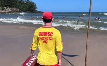 Bombeiros realizam 210 mil ações preventivas nas praias Costas de um guarda-vida civil usando camiseta amarela com a sigla "CBMSC" e bermuda vermelha. Ele segura um flutuador de resgate (lifebelt) vermelho e observa o mar agitado com banhistas ao fundo. Ilustra as 210 mil ações preventivas no litoral sul.