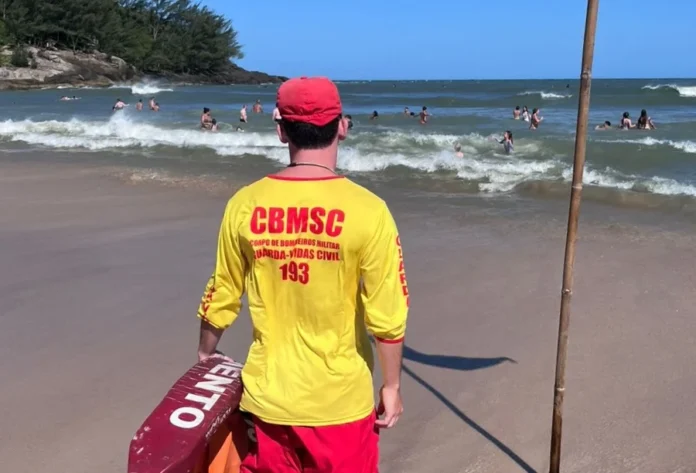 Bombeiros realizam 210 mil ações preventivas nas praias Costas de um guarda-vida civil usando camiseta amarela com a sigla "CBMSC" e bermuda vermelha. Ele segura um flutuador de resgate (lifebelt) vermelho e observa o mar agitado com banhistas ao fundo. Ilustra as 210 mil ações preventivas no litoral sul.