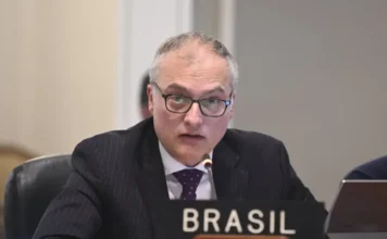 Brasil condena ação dos EUA e vê afronta à soberania Close-up do embaixador Benoni Belli, usando óculos e terno risca-de-giz, falando ao microfone durante sessão na OEA. À sua frente, uma placa com a inscrição "BRASIL".