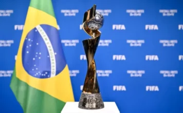 Brasil cria regras para a Copa do Mundo Feminina 2027 Fotografia em destaque do troféu oficial da Copa do Mundo Feminina da Fifa, uma peça dourada e prateada com design espiralado que envolve uma bola de futebol no topo. Ao fundo, de forma levemente desfocada, aparecem a bandeira do Brasil e um painel azul com o logotipo da Fifa, simbolizando o regime jurídico de proteção especial e os direitos de propriedade intelectual estabelecidos pela Medida Provisória 1335/26 para o mundial no país.