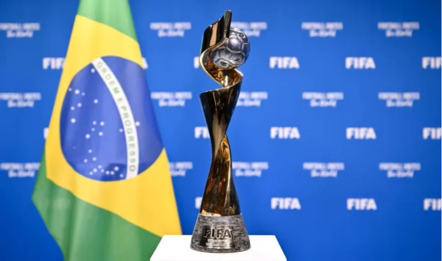 Fotografia em destaque do troféu oficial da Copa do Mundo Feminina da Fifa, uma peça dourada e prateada com design espiralado que envolve uma bola de futebol no topo. Ao fundo, de forma levemente desfocada, aparecem a bandeira do Brasil e um painel azul com o logotipo da Fifa, simbolizando o regime jurídico de proteção especial e os direitos de propriedade intelectual estabelecidos pela Medida Provisória 1335/26 para o mundial no país.