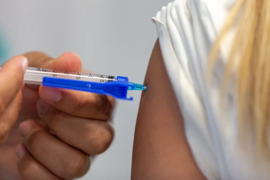 Close-up de um profissional de saúde aplicando uma vacina no braço de uma pessoa vestindo camiseta branca, utilizando uma seringa com detalhe em azul.