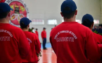 CBMSC abre concurso com salários de até R$ 21,2 mil Fotografia em plano médio mostrando as costas de três militares vestindo agasalhos vermelhos e bonés azuis. No destaque central, a estampa em letras brancas no uniforme diz: "CURSO DE FORMAÇÃO DE OFICIAIS - CADETE". Ao fundo, é possível ver o emblema do Corpo de Bombeiros Militar de Santa Catarina em uma parede e outros militares em formação, reforçando o ambiente institucional da corporação.