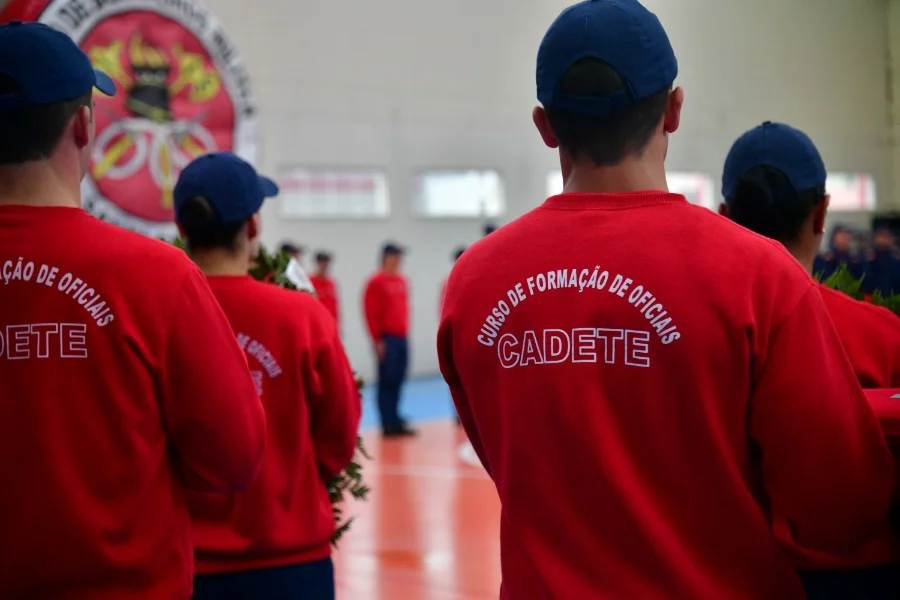 Fotografia em plano médio mostrando as costas de três militares vestindo agasalhos vermelhos e bonés azuis. No destaque central, a estampa em letras brancas no uniforme diz: 