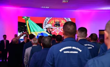 CBMSC inicia comemorações de 100 anos com lançamentos Bombeiros militares de Santa Catarina de costas, vestindo fardas azuis com a inscrição "Bombeiro Militar Santa Catarina", acompanham a solenidade de abertura dos 100 anos da corporação em Florianópolis. Ao fundo, uma tela exibe a bandeira do Brasil e o brasão do CBMSC sob iluminação cênica.
