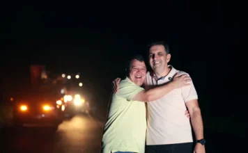 CEPRAG conclui modernização elétrica na Serra do Faxinal Fotografia noturna na Serra do Faxinal mostra o prefeito de Praia Grande e o presidente da CEPRAG, Patrique Alencar Homem, celebrando a conclusão da obra. Eles estão abraçados e sorridentes no centro da estrada, simbolizando a parceria entre as instituições. Ao fundo, um caminhão de serviço com faróis e sinalizadores acesos ilumina a via recém-modernizada.