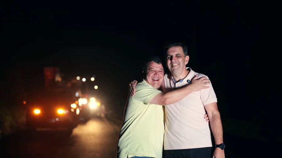 Fotografia noturna na Serra do Faxinal mostra o prefeito de Praia Grande e o presidente da CEPRAG, Patrique Alencar Homem, celebrando a conclusão da obra. Eles estão abraçados e sorridentes no centro da estrada, simbolizando a parceria entre as instituições. Ao fundo, um caminhão de serviço com faróis e sinalizadores acesos ilumina a via recém-modernizada.