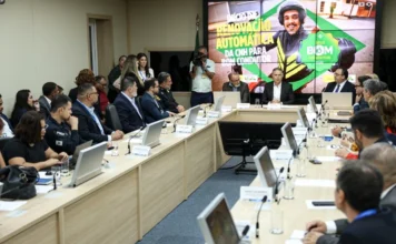 CNH automática: governo libera 1º lote para bons condutores Foto de uma reunião em uma sala de conferências, onde o Ministério dos Transportes anuncia o início da renovação automática da Carteira Nacional de Habilitação (CNH) para bons condutores. Um grande telão ao fundo exibe a frase "INÍCIO DE RENOVAÇÃO AUTOMÁTICA DA CNH PARA BOM CONDUTOR" e o logo do "SELO BOM CONDUTOR". Vários oficiais e participantes estão sentados ao redor de uma longa mesa, acompanhando a apresentação.
