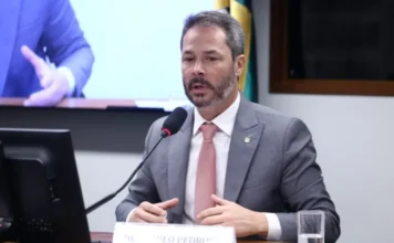 Câmara aprova transparência em taxa de iluminação pública O deputado Saulo Pedroso (PSD-SP), relator da proposta sobre a Cosip, fala ao microfone durante uma comissão na Câmara dos Deputados. Ele veste terno cinza e gravata rosa, e gesticula enquanto defende a transparência e a modernização da iluminação pública.