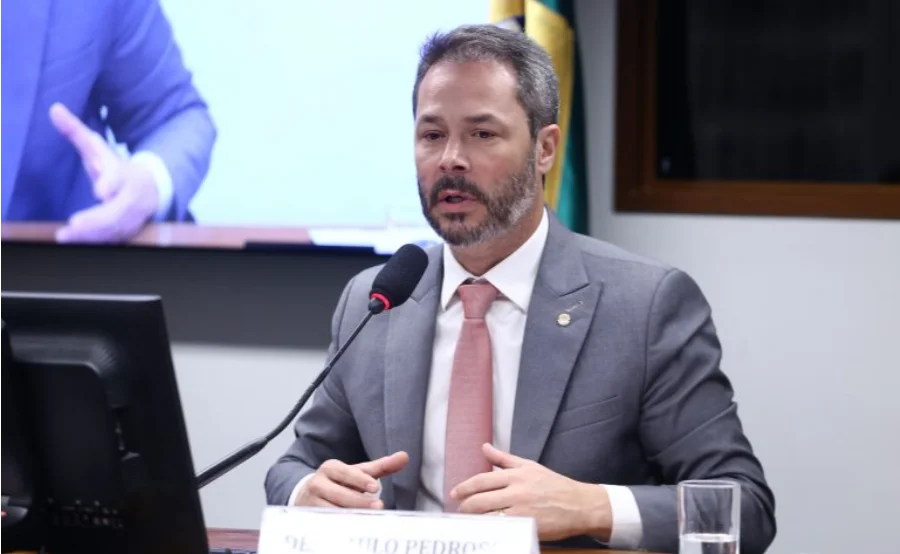 O deputado Saulo Pedroso (PSD-SP), relator da proposta sobre a Cosip, fala ao microfone durante uma comissão na Câmara dos Deputados. Ele veste terno cinza e gravata rosa, e gesticula enquanto defende a transparência e a modernização da iluminação pública.