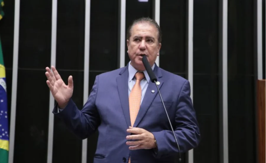Câmara propõe fim de multas duplicadas para empresas O deputado Jonas Donizette, vestindo terno azul e gravata laranja, discursa em um púlpito da Câmara dos Deputados com a bandeira do Brasil ao fundo.