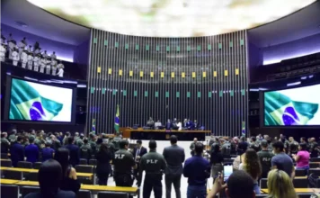 Câmara publica edital para Policial Legislativo Federal Fotografia do Plenário da Câmara dos Deputados em Brasília durante uma cerimônia oficial. No centro, diversos Policiais Legislativos Federais (PLF) estão de pé, de costas para a câmera, vestindo seus uniformes operacionais verdes com a sigla "PLF" em destaque nas costas. Ao fundo, a Mesa Diretora é composta por autoridades, ladeada por grandes telões que exibem a bandeira do Brasil. O ambiente solene ilustra a corporação para a qual o novo edital oferece 40 vagas imediatas e salário inicial superior a R$ 21 mil.