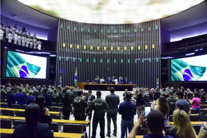 Câmara publica edital para Policial Legislativo Federal Fotografia do Plenário da Câmara dos Deputados em Brasília durante uma cerimônia oficial. No centro, diversos Policiais Legislativos Federais (PLF) estão de pé, de costas para a câmera, vestindo seus uniformes operacionais verdes com a sigla "PLF" em destaque nas costas. Ao fundo, a Mesa Diretora é composta por autoridades, ladeada por grandes telões que exibem a bandeira do Brasil. O ambiente solene ilustra a corporação para a qual o novo edital oferece 40 vagas imediatas e salário inicial superior a R$ 21 mil.