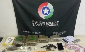 Casal é preso com drogas, arma e R$ 5 mil no Sul do Estado Materiais apreendidos pela Polícia Militar dispostos sobre uma mesa: tabletes de maconha, porções de cocaína, um revólver com munições, duas balanças de precisão, um celular e diversas cédulas de dinheiro em espécie. Ao fundo, vê-se a bandeira preta com o brasão da Polícia Militar de Santa Catarina.