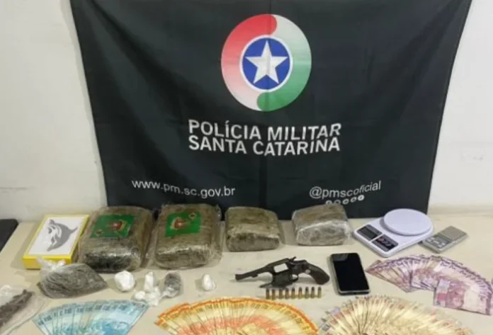 Casal é preso com drogas, arma e R$ 5 mil no Sul do Estado Materiais apreendidos pela Polícia Militar dispostos sobre uma mesa: tabletes de maconha, porções de cocaína, um revólver com munições, duas balanças de precisão, um celular e diversas cédulas de dinheiro em espécie. Ao fundo, vê-se a bandeira preta com o brasão da Polícia Militar de Santa Catarina.