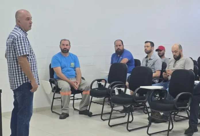 Celesc e Prefeitura ampliam Limpa Fios em Criciúma Fotografia mostra uma reunião técnica em uma sala de conferências, onde um homem em pé, vestindo camisa xadrez, fala para um grupo de profissionais sentados em semicírculo. Entre os participantes, nota-se a presença de técnicos com uniformes da Celesc e de empresas de telecomunicações, evidenciando o alinhamento estratégico para as ações do programa Limpa Fios em Criciúma.