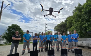 Celesc investe R$ 950 mil em drones para prevenir falhas Uma equipe de profissionais da Celesc, usando camisetas azuis, posa em uma área externa com torres de transmissão de energia ao fundo. No chão, à frente do grupo, estão várias maletas de transporte pretas abertas e dois drones pousados. Um terceiro drone de grande porte está voando pairado no ar, logo acima da equipe.
