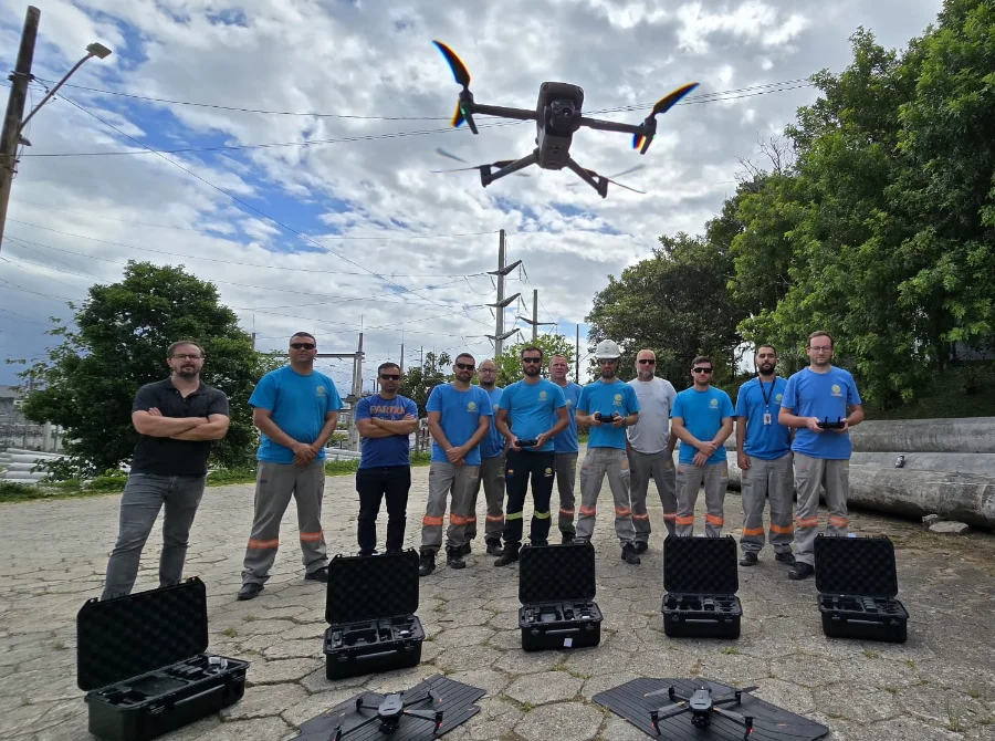Uma equipe de profissionais da Celesc, usando camisetas azuis, posa em uma área externa com torres de transmissão de energia ao fundo. No chão, à frente do grupo, estão várias maletas de transporte pretas abertas e dois drones pousados. Um terceiro drone de grande porte está voando pairado no ar, logo acima da equipe.